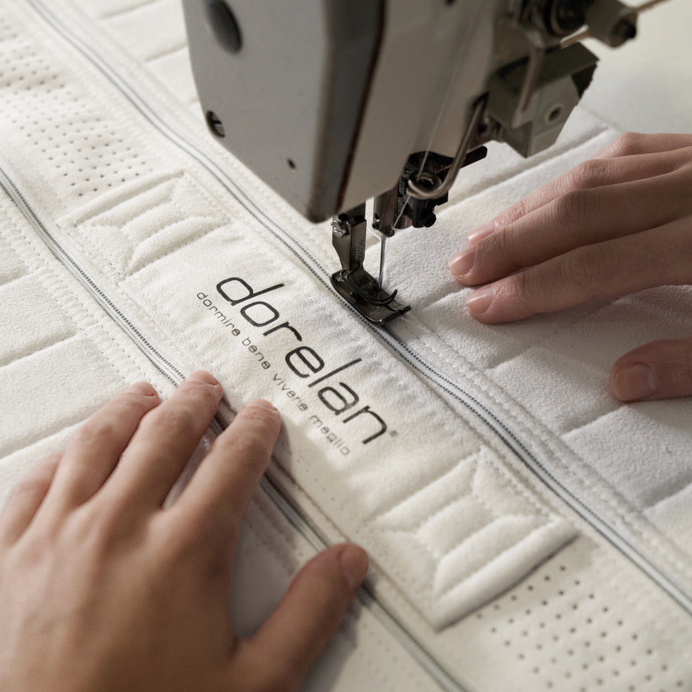 Primo piano delle mani di un artigiano che cucisce con cura l'etichetta di Dorelan su un materasso, evidenziando l'alta qualità e l'attenzione al dettaglio nella produzione.